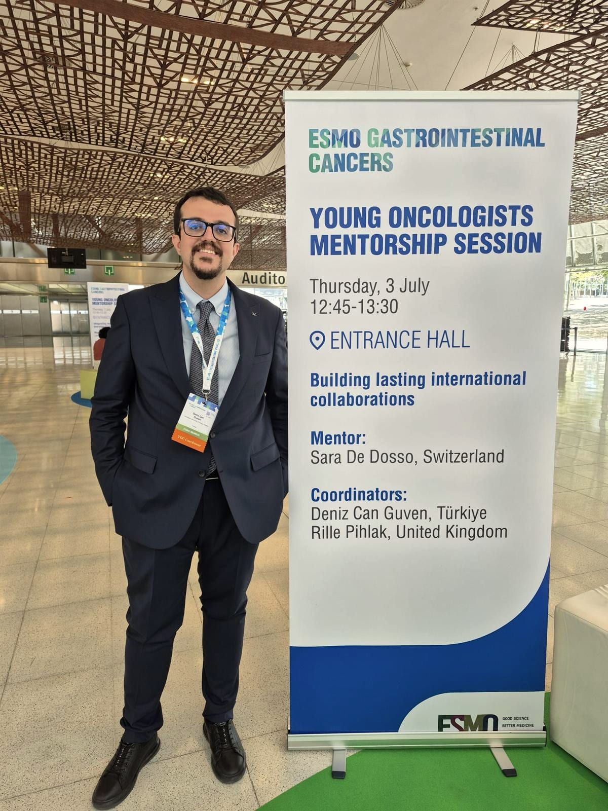 Doç. Dr. Deniz Can Güven, ESMO Gastrointestinal Kanserler Kongresi'nde koordinatörü olduğu Genç Onkologlar Mentorluk Oturumu afişinin yanında