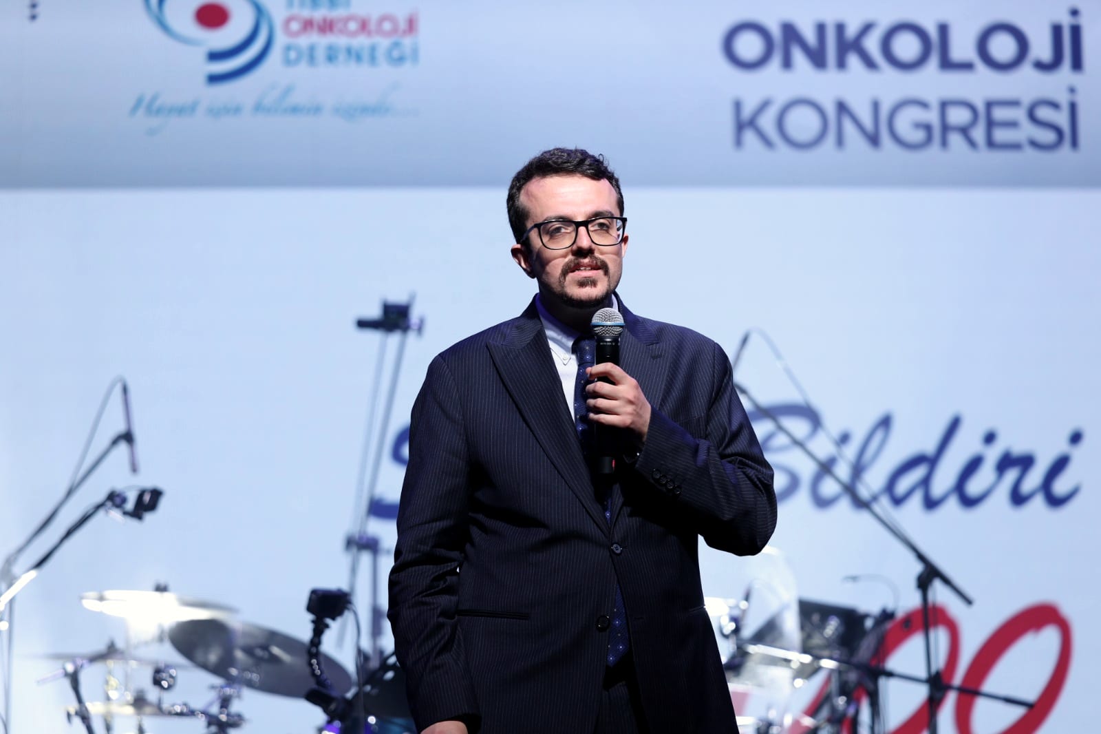 Doç. Dr. Deniz Can Güven, Türk Tıbbi Onkoloji Kongresi sahnesinde mikrofonla konuşurken