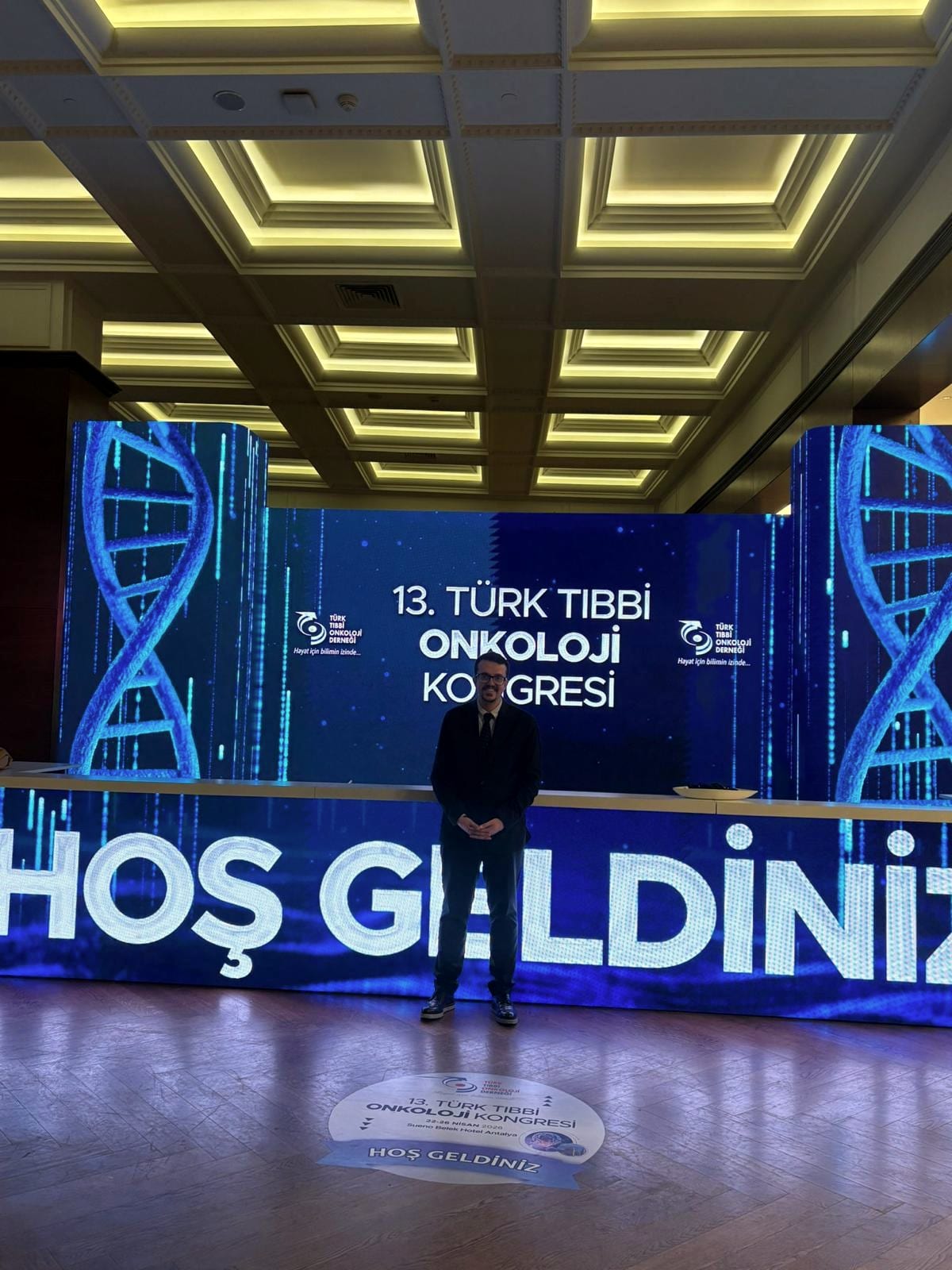 Doç. Dr. Deniz Can Güven, DNA sarmalı görselleriyle aydınlatılmış kongre sahnesinin önünde